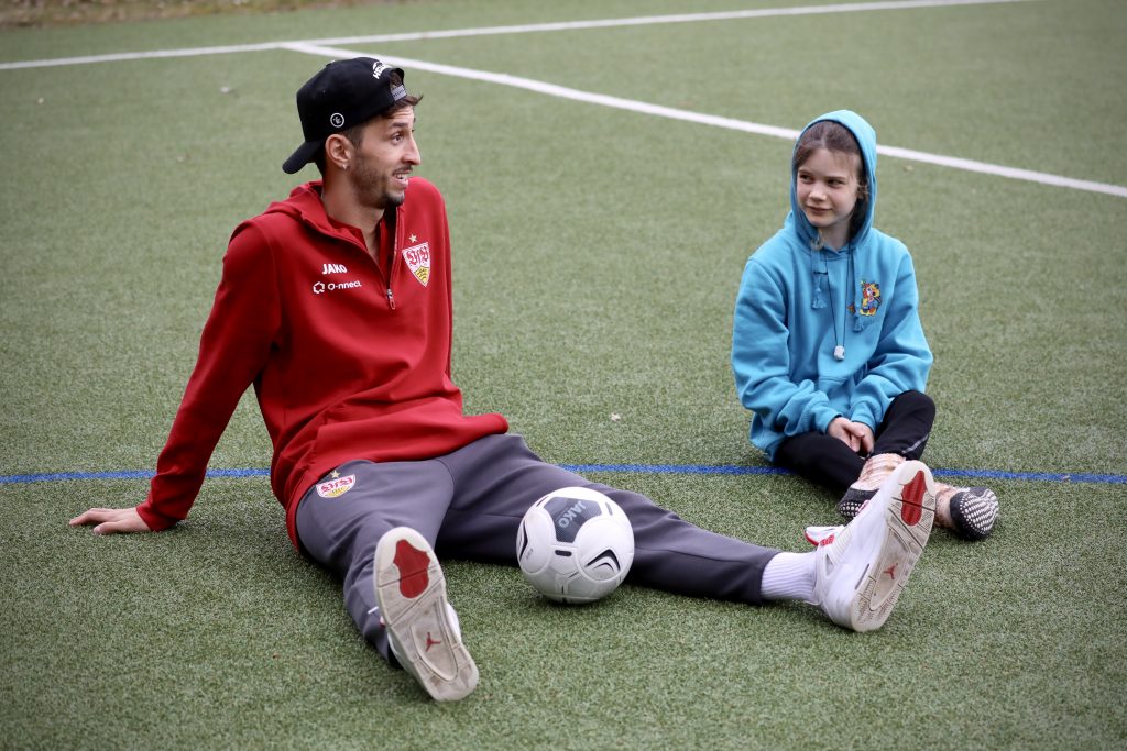 VfB-Profi Atakan Karazor sitzt auf dem Fußballplatz der Nachsorgeklinik Tannheim neben einer kleinen Patientin und plaudert mit ihr